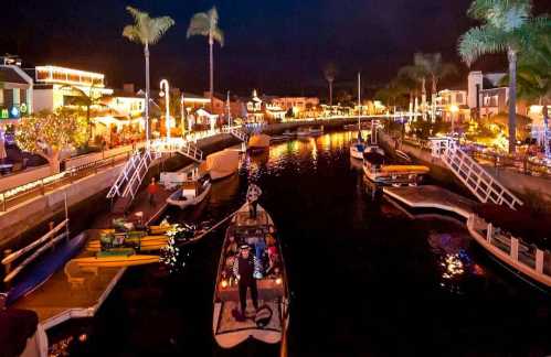 A scenic nighttime view of a canal lined with illuminated buildings and boats, surrounded by palm trees.