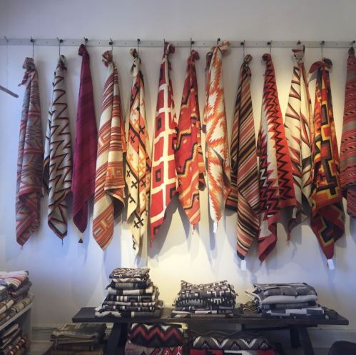 A display of colorful woven textiles hanging on a wall, with folded blankets stacked below.
