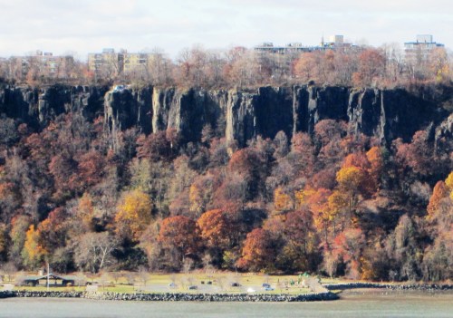 A rocky cliff with autumn foliage overlooks a park and water, with buildings visible in the background.