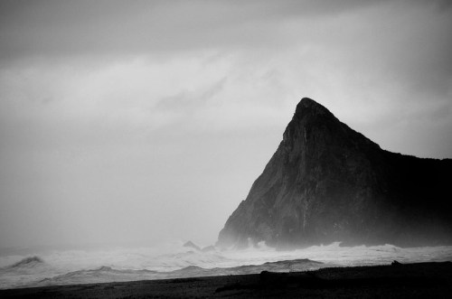 A dramatic black and white image of a tall, jagged rock formation by a stormy ocean. Waves crash against the shore.