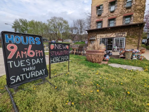 A coffee shop with a sign displaying hours and a new strawberry horchata, surrounded by greenery and flowers.
