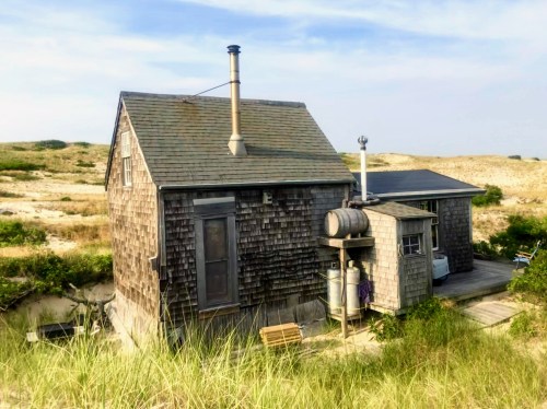 A rustic wooden house with a chimney, surrounded by grassy dunes under a clear sky.