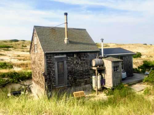 A rustic wooden house with a chimney, surrounded by grassy dunes under a clear sky.