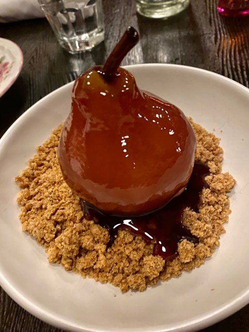 A glossy caramelized pear sits atop a bed of crumbled cookie, drizzled with sauce on a white plate.