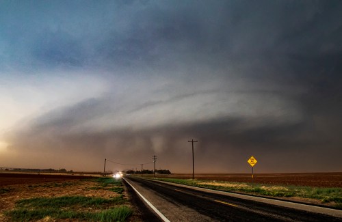 A dramatic storm cloud looms over a rural road, with dark skies and distant lightning illuminating the scene.