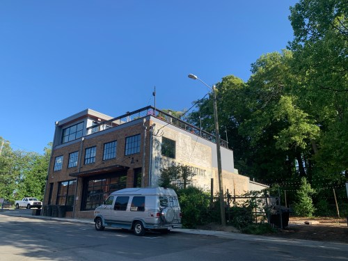 A brick building with large windows and a rooftop, surrounded by trees and a street with a parked van. Clear blue sky.