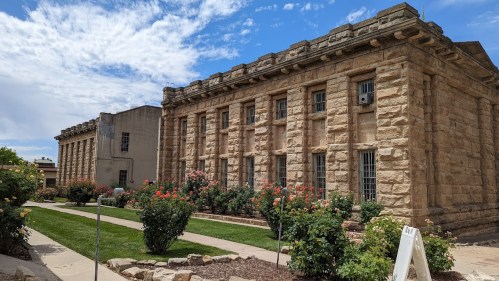 Historic stone buildings with large windows, surrounded by green lawns and blooming rose bushes under a partly cloudy sky.