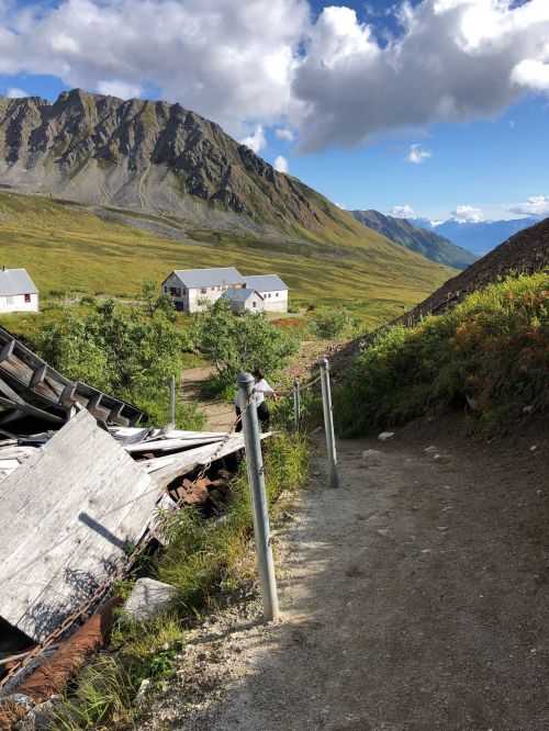 A dirt path leads through a mountainous landscape with scattered houses and lush greenery under a blue sky.