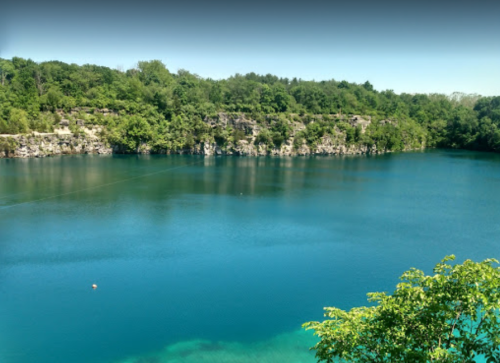 A serene blue lake surrounded by lush green trees and rocky cliffs under a clear sky.