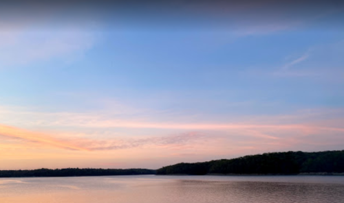 A serene lake at sunset, with soft pink and blue hues in the sky and a forested shoreline in the distance.