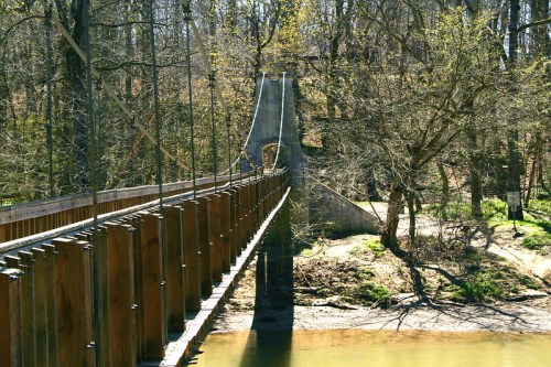A wooden suspension bridge spans a river, surrounded by trees and greenery in a serene natural setting.
