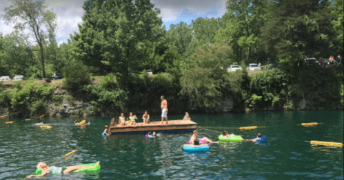 People swimming and relaxing on inflatable floats in a green lake, with a wooden platform and trees in the background.