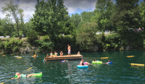 A sunny day at a swimming area with people on a dock and floating in colorful inner tubes on a green lake.