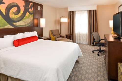 A cozy hotel room featuring a large bed, armchair, desk, and a window with curtains. Modern decor and warm colors.