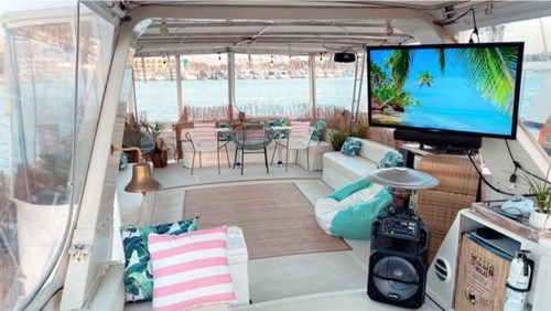 Cozy boat interior with seating, a TV displaying a beach scene, and views of water and city in the background.