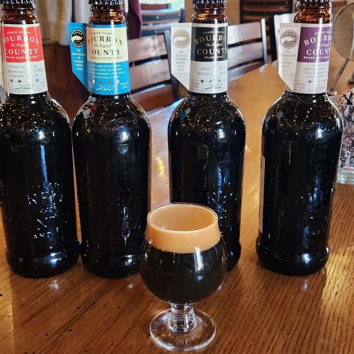 Four bottles of Bourbon County beer lined up on a wooden table, with a small glass of dark beer in the center.