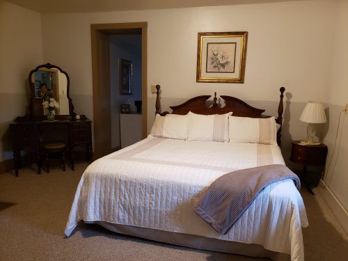 A cozy bedroom featuring a large bed, a wooden dresser, and soft lighting, with neutral-colored walls and carpet.