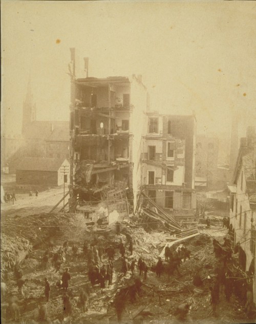 Historic photo of a building collapse, with debris scattered and people gathered around the site.