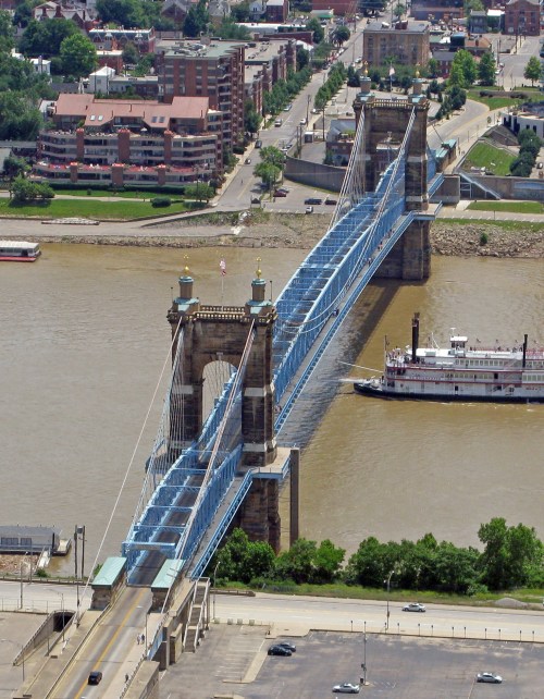 Aerial view of a blue suspension bridge over a river, with a steamboat passing underneath and buildings in the background.