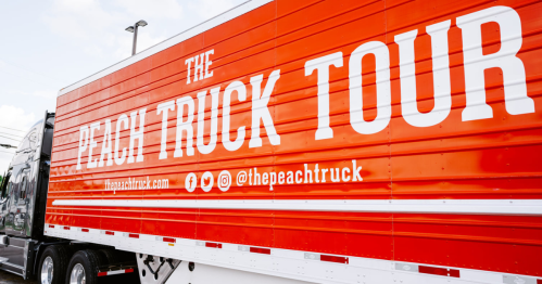 A bright orange truck with "The Peach Truck Tour" written on the side, parked in a lot.