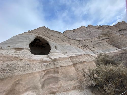 A rocky cliff with a large hole, surrounded by layered sediment and sparse vegetation under a cloudy sky.