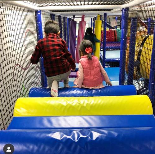 Two children climb colorful foam obstacles in a play area, with netting and soft surfaces surrounding them.
