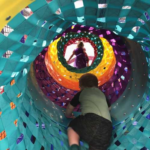 A child crawls through a colorful tunnel, with another child visible at the far end, surrounded by vibrant patterns.