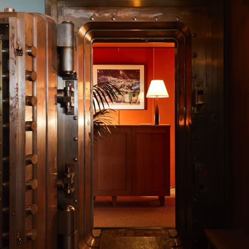 A vintage bank vault door opens to a warmly lit room with a lamp, artwork, and a plant.