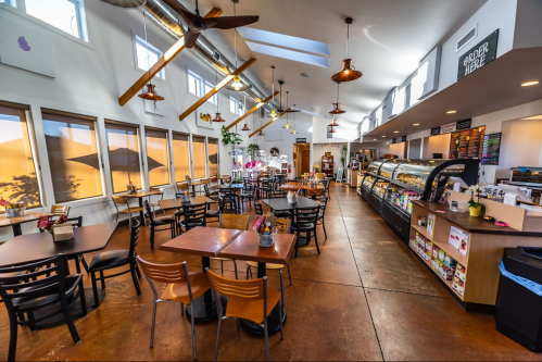 Bright café interior with wooden tables, chairs, and a display case, featuring large windows and modern lighting.