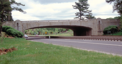 A concrete bridge arches over a highway, surrounded by greenery and trees under a cloudy sky.
