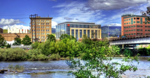Cityscape featuring buildings along a river, with greenery in the foreground and a blue sky with clouds above.