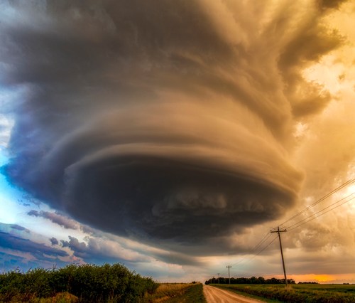 A dramatic, swirling storm cloud looms over a rural road, illuminated by a warm sunset. Power lines stretch in the background.