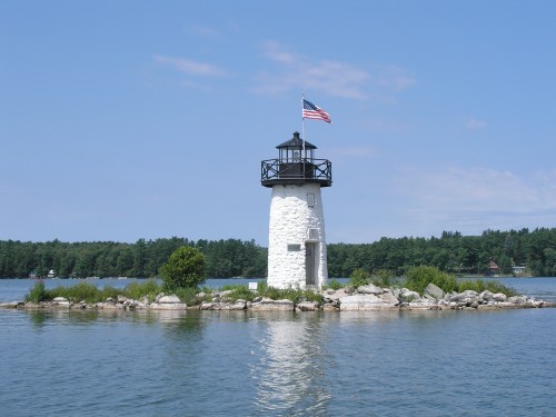 A white lighthouse with a black top and an American flag stands on a small island surrounded by calm water and greenery.