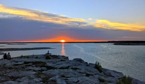 A serene sunset over a calm lake, with silhouettes of people on rocky shores and vibrant colors in the sky.