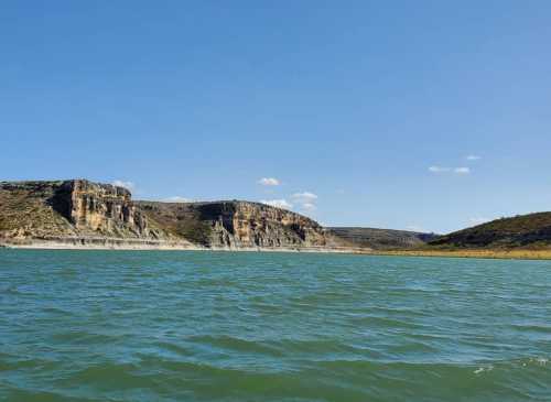 A serene lake surrounded by rocky cliffs and green hills under a clear blue sky.