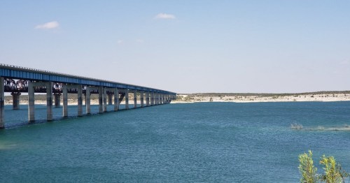 A long bridge spans over a calm blue lake, with clear skies and distant land visible in the background.