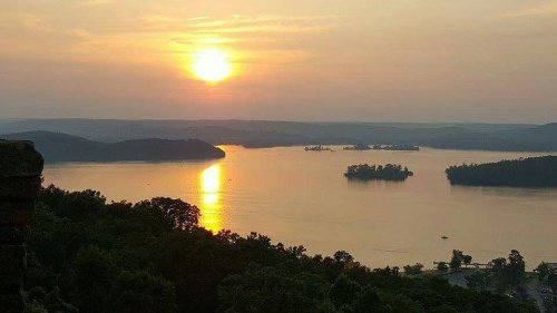 A serene sunset over a calm lake, with silhouettes of distant hills and islands reflecting on the water.