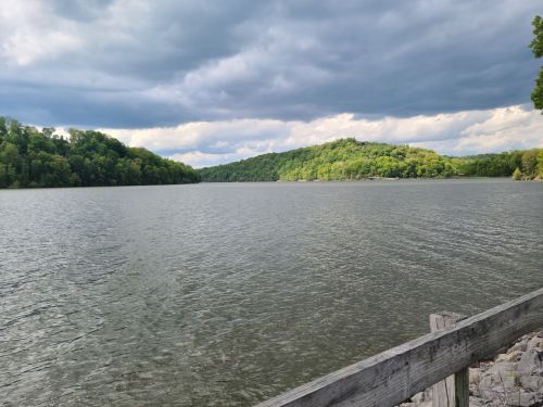 A serene lake surrounded by green hills under a cloudy sky, with a wooden railing in the foreground.
