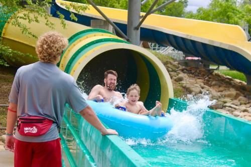 A man and a child joyfully ride a water slide in an inflatable tube, splashing water as they descend. A lifeguard watches nearby.
