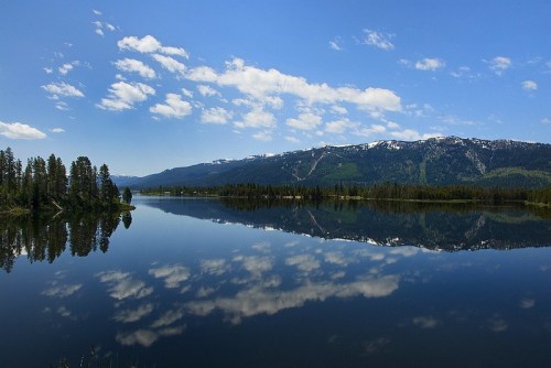 A serene lake reflects a clear blue sky and mountains, surrounded by lush greenery and scattered clouds.