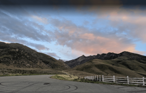 A winding road curves through green mountains under a colorful sky with clouds at sunset. A white fence lines the scene.