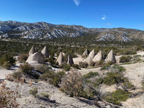 A desert landscape featuring conical rock formations surrounded by sparse vegetation and distant mountains under a blue sky.
