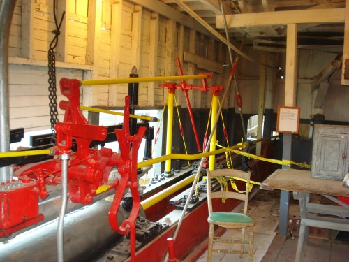Interior of a historic boat, featuring red and yellow machinery, wooden walls, and a chair in a rustic setting.