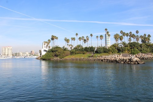 A serene waterfront view featuring palm trees, a rocky shore, and boats in the distance under a clear blue sky.