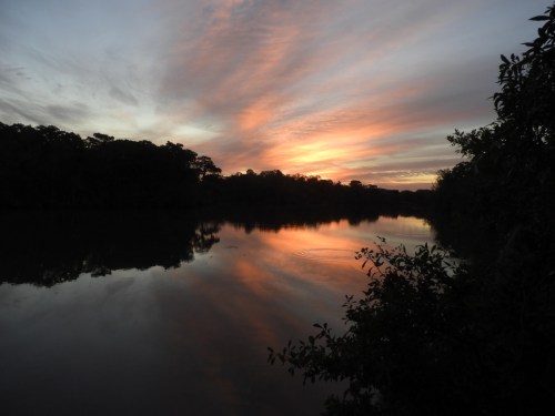A serene sunset over a calm river, with colorful clouds reflecting on the water and trees lining the shore.