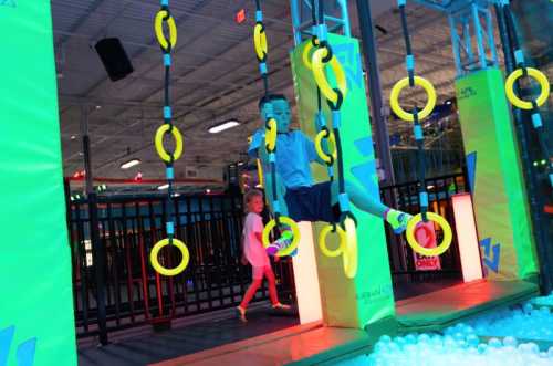 A child swings on bright yellow rings in a colorful indoor play area, with another child in the background.