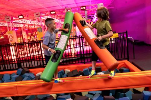 Two children play on a balance beam, using colorful foam sticks in a vibrant indoor trampoline park.