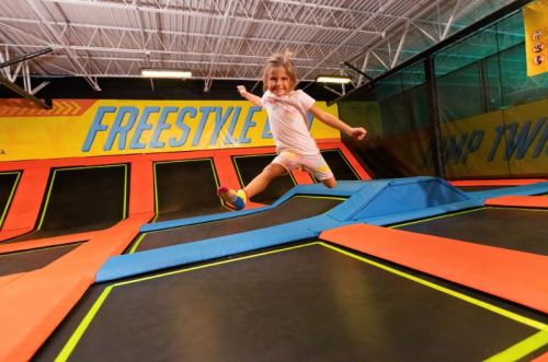 A young girl joyfully jumps on a trampoline at a trampoline park, surrounded by colorful mats and walls.