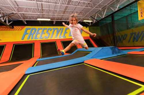 A young girl joyfully jumps on a trampoline at a trampoline park, surrounded by colorful mats and walls.