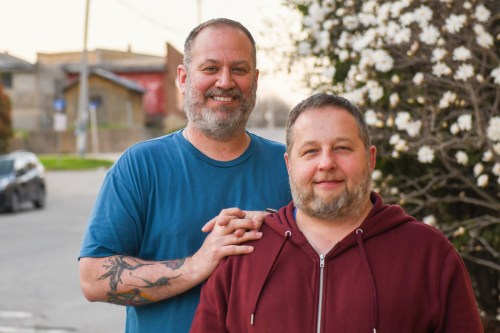 Two men stand outdoors, smiling, with one in a blue shirt and the other in a maroon hoodie, surrounded by blooming flowers.